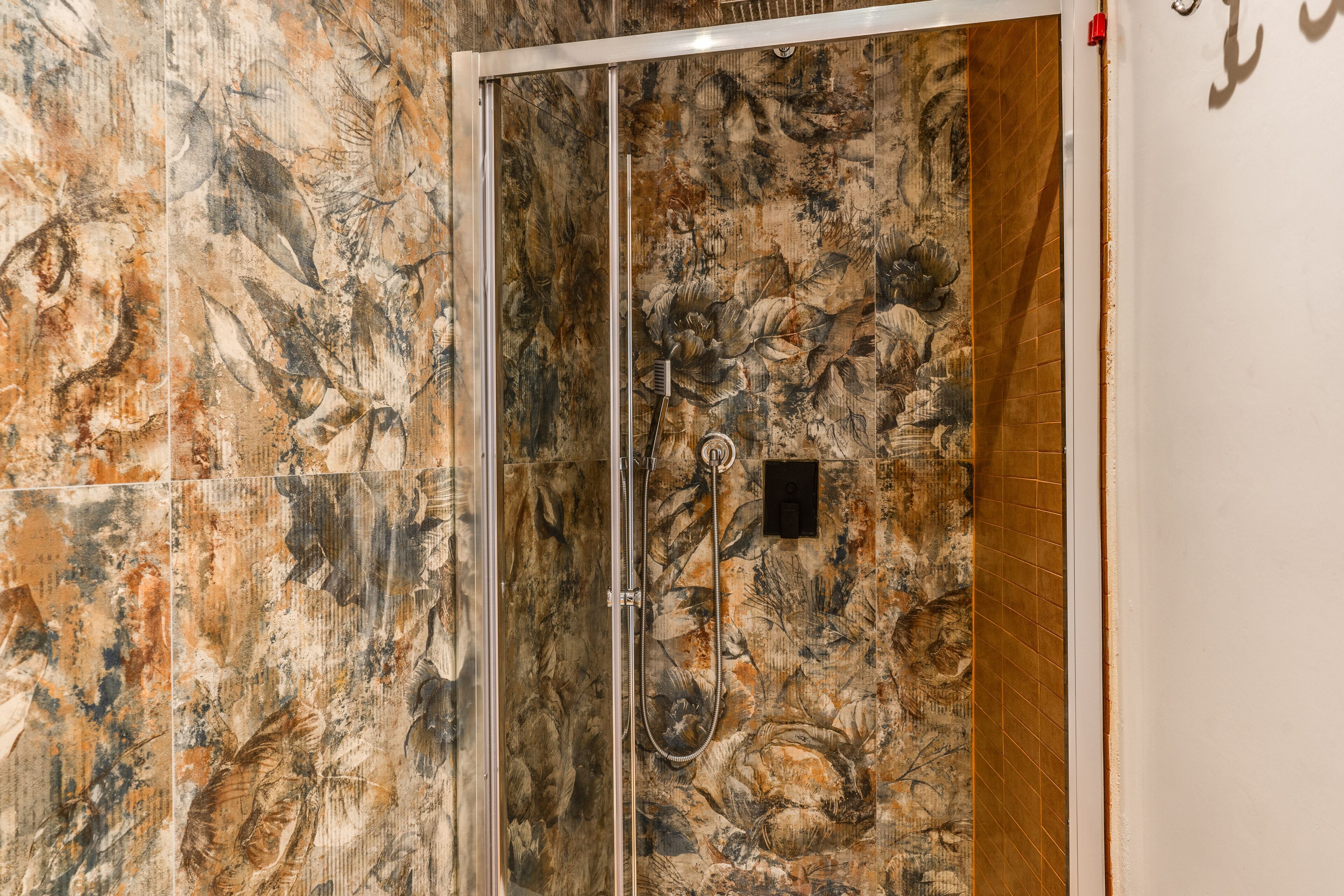 A shower area with a glass door, a hand shower, and a wall with a floral tile pattern.