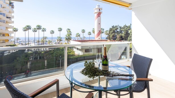 En balkon med et glasbord, en champagneflaske og en stol. Et fyrtårn er synligt i baggrunden.