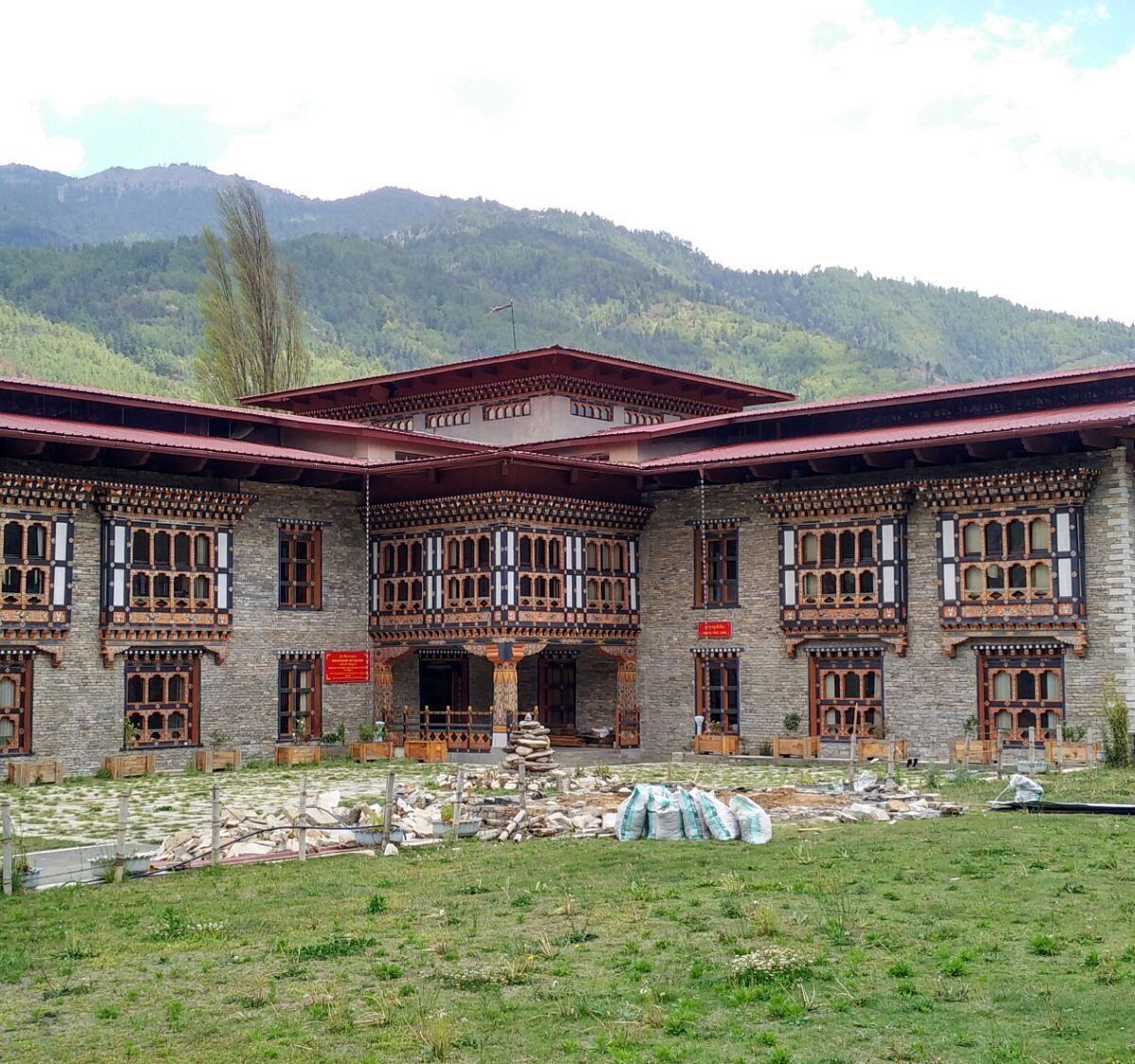 A traditional building with multiple windows and a red roof, surrounded by a grassy area and mountains in the background.