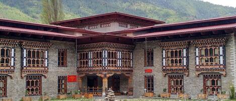 A traditional building with multiple windows and a red roof, surrounded by a grassy area and mountains in the background.