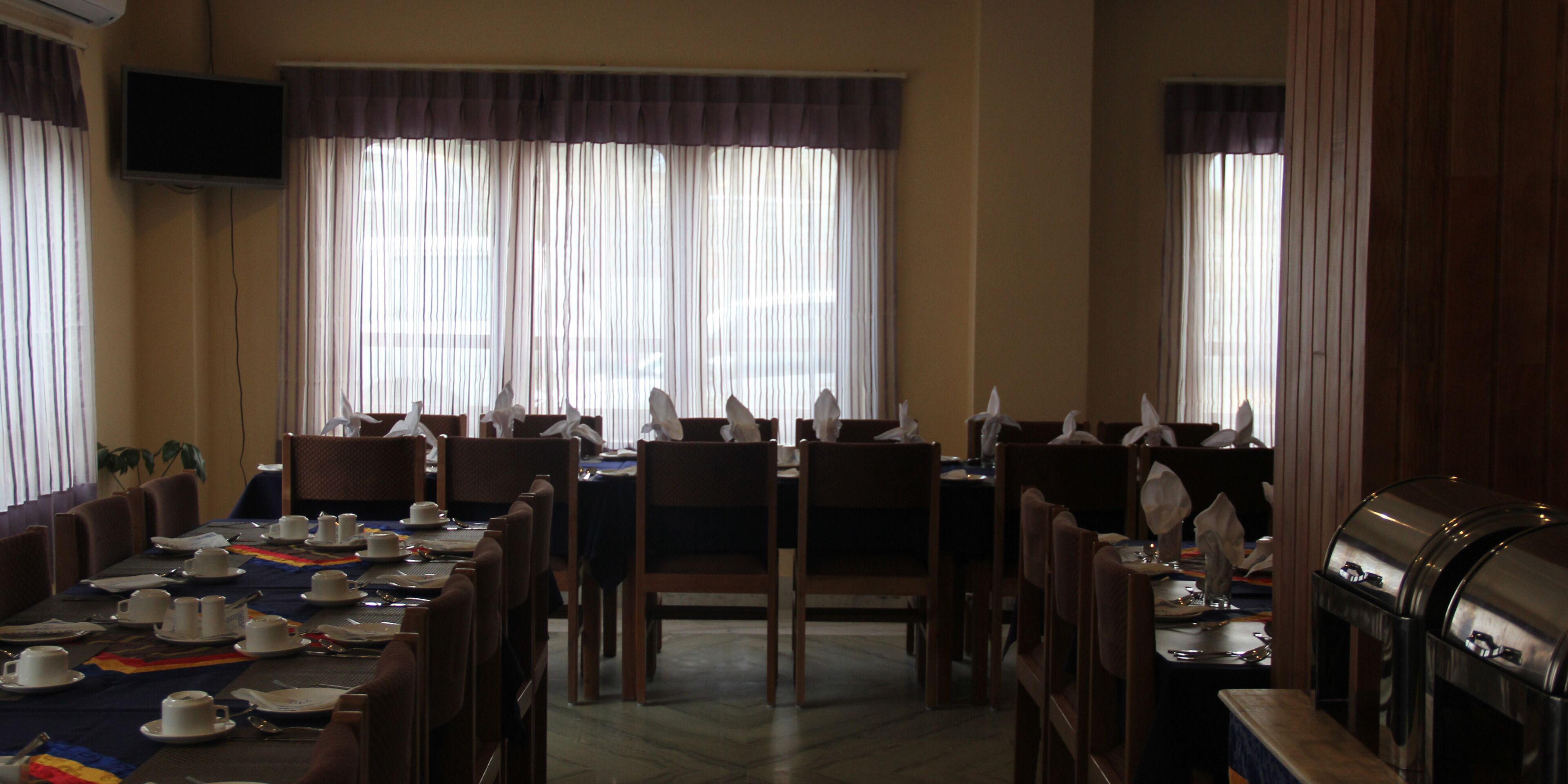Un comedor con mesas dispuestas para una comida, sillas, un televisor montado en la pared y ventanas amplias con cortinas.