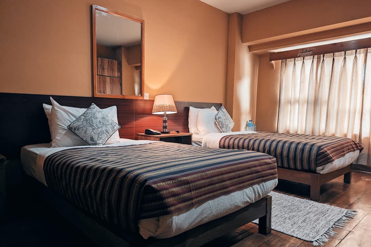 A hotel room with two beds, a nightstand, a lamp, a mirror, and a window with curtains.