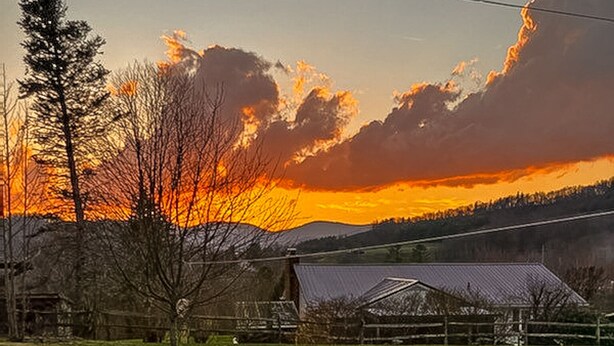 Eine Terrasse mit zwei Adirondack-Stühlen und einer Feuerstelle, vor einer ländlichen Landschaft bei Sonnenuntergang.