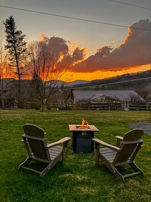 Un patio con dos sillas Adirondack y un brasero, con el atardecer como fondo sobre un paisaje rural.