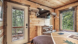 A wooden cabin bedroom with a large window, a bed with a patterned blanket, and a wooden bench with logs. - NEXT TO DOLLYWOOD! Treetop Cabin 2br/2ba, Private Hot Tub, Gas Grill, 2 Decks (Pigeon Forge)