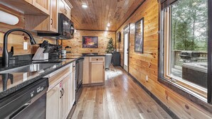 A wooden cabin interior with a kitchen area, wooden cabinets, a dishwasher, and large windows offering a view of the outdoors. - NEXT TO DOLLYWOOD! Treetop Cabin 2br/2ba, Private Hot Tub, Gas Grill, 2 Decks (Pigeon Forge)