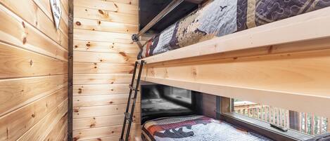 A cabin with wooden walls, a bunk bed, a ladder, and a window with a view.
