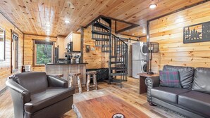 A cabin interior with a wooden interior, a sofa, a coffee table, a dining table, and a kitchen area with a spiral staircase. - NEXT TO DOLLYWOOD! Treetop Cabin 2br/2ba, Private Hot Tub, Gas Grill, 2 Decks (Pigeon Forge)