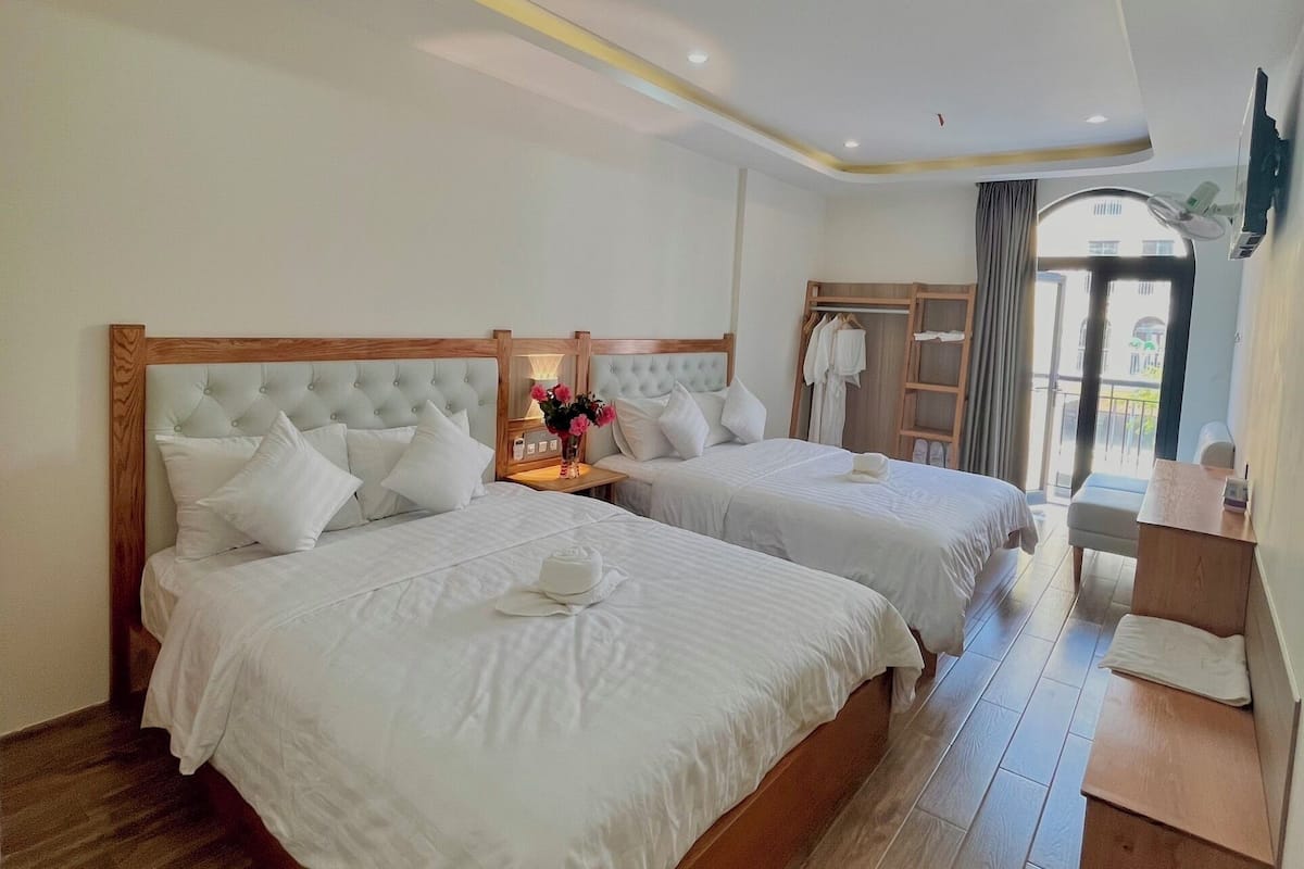 A hotel room with two beds, a wooden headboard, a bedside table with a vase of flowers, and a view of the outside through a large window.