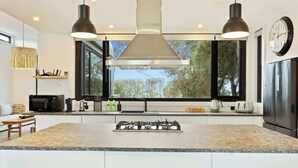 Een moderne keuken met een centraal eiland, roestvrijstalen apparatuur, hangende lampen en een groot raam met uitzicht op bomen.