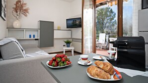 A modern living room with a sofa, a dining table with strawberries and croissants, a coffee machine, and a TV on the wall.