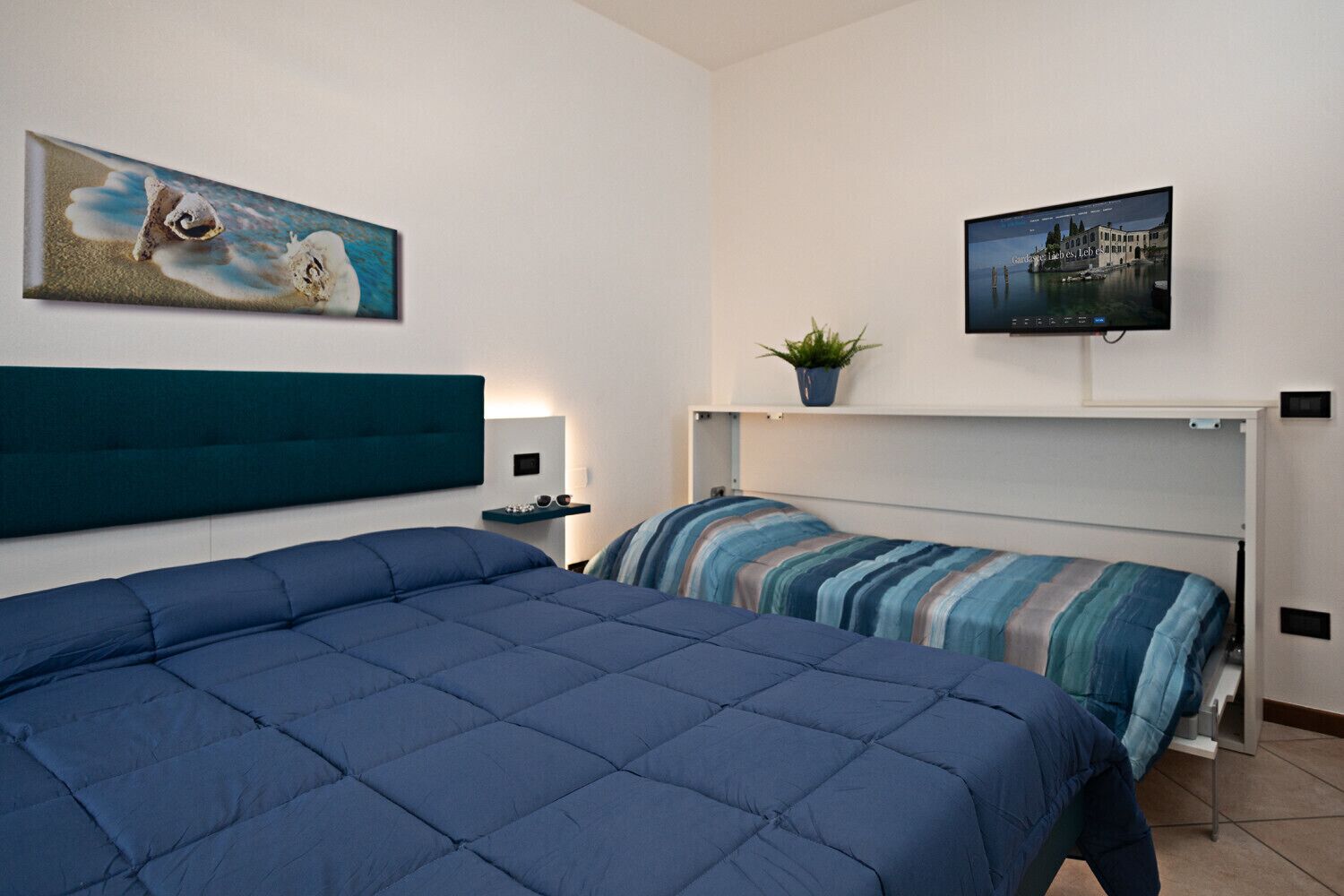 Une chambre avec deux lits, une tête de lit, une table de chevet avec une lampe, une télévision et une étagère murale agrémentée d’une plante.