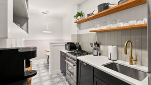 A modern kitchen with a gas stove, oven, and coffee maker. There are wooden shelves above the sink with glassware and a plant.