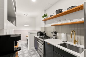 A modern kitchen with a gas stove, oven, and coffee maker. There are wooden shelves above the sink with glassware and a plant.
