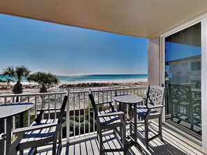 A balcony with a view of the beach, featuring wooden chairs and a table.