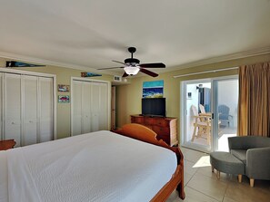 A bedroom with a bed, a ceiling fan, a TV, a sliding glass door to a balcony, and a chair.