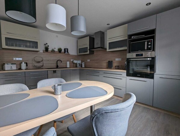 A modern kitchen with grey cabinetry, a wooden dining table with grey cushions, and built-in appliances including a microwave and oven. - Modern semi-detached house with large garden (Lemgo)
