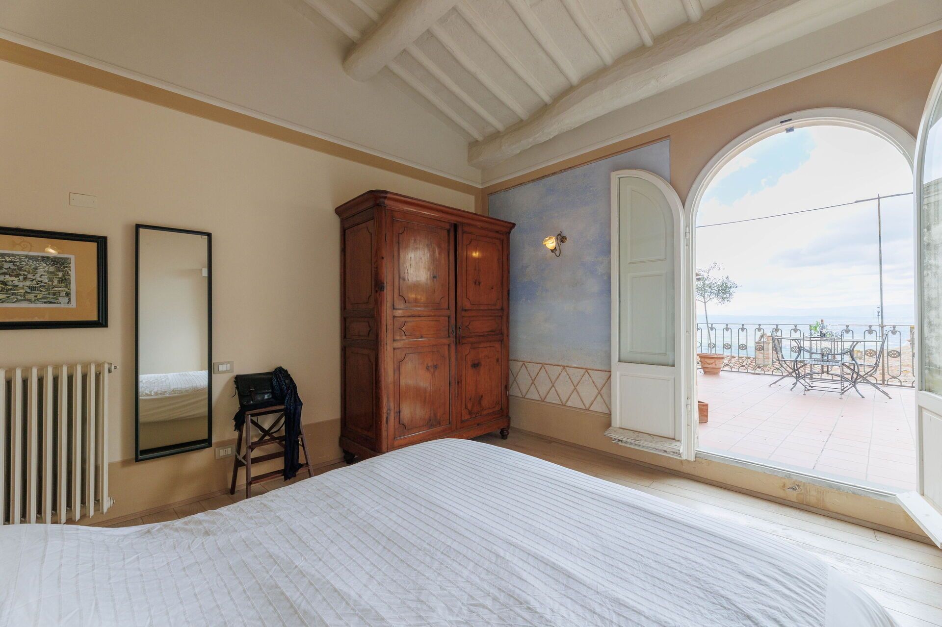 Een slaapkamer met een groot bed, een houten garderobe, een radiator, een spiegel en uitzicht op een balkon met buitenmeubilair.