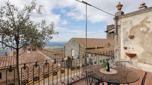A terrace with a round metal table and chairs, a view of the town below, and a stone building with a tiled roof.