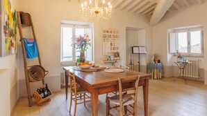 A spacious dining area with a wooden table, chairs, and a chandelier. There is a window with a view, a radiator, and a shelf with a vase and picture frame.