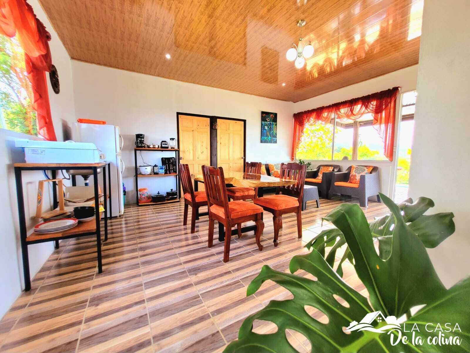 A spacious living area with a wooden ceiling, a dining table with chairs, a kitchen area with appliances, and a large window with curtains.