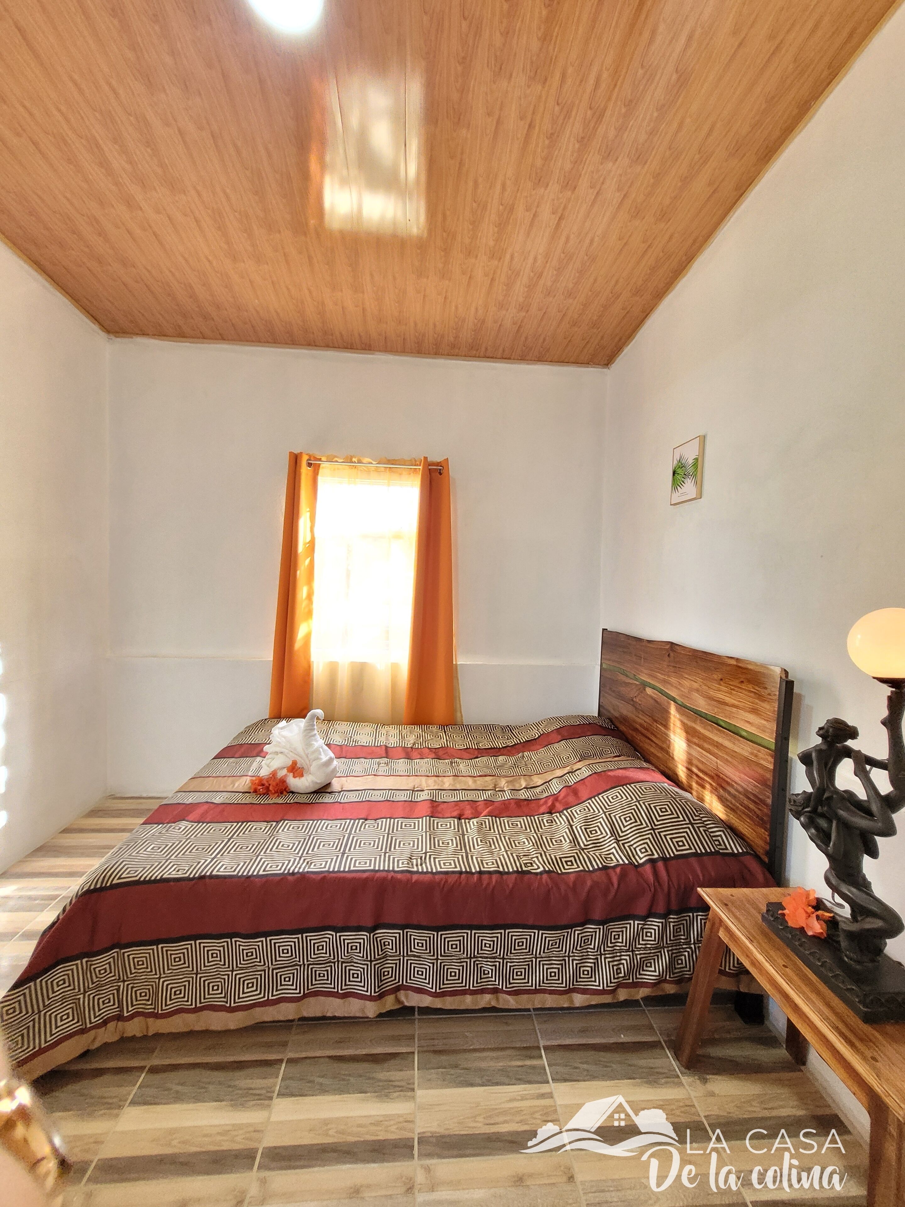 A bedroom with a wooden ceiling, a bed with patterned bedding, a window with orange curtains, and a small table with a lamp.