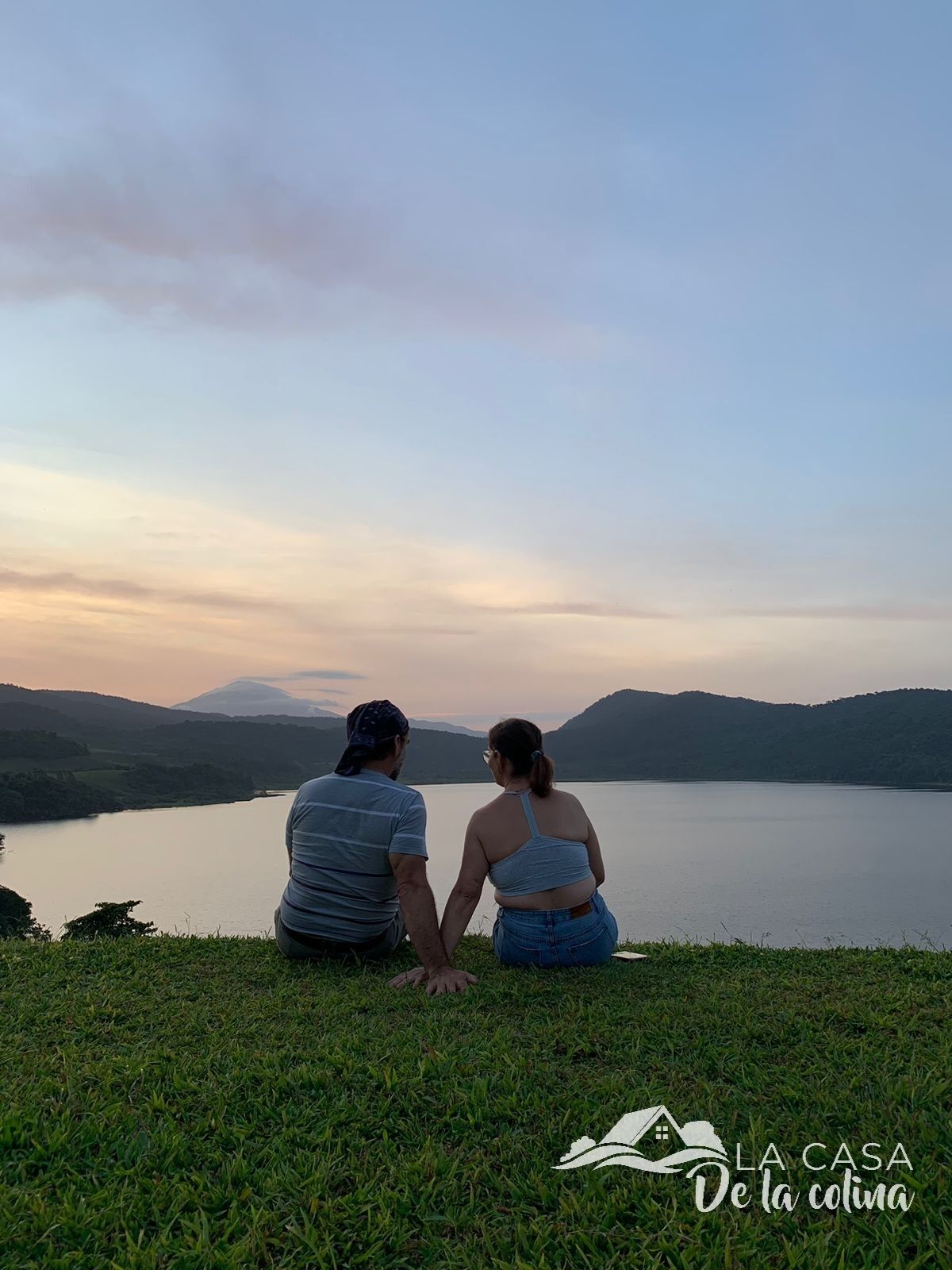 Deux personnes assises sur l’herbe, face à un lac, avec des montagnes en arrière-plan.