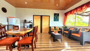 Un vaste espace de vie comprenant une table à manger en bois, des chaises, un canapé et une table basse. Il y a également un coin cuisine équipé d’appareils électroménagers et d’un réfrigérateur. La pièce est dotée d’un sol carrelé et d’une grande fenêtre avec des rideaux.