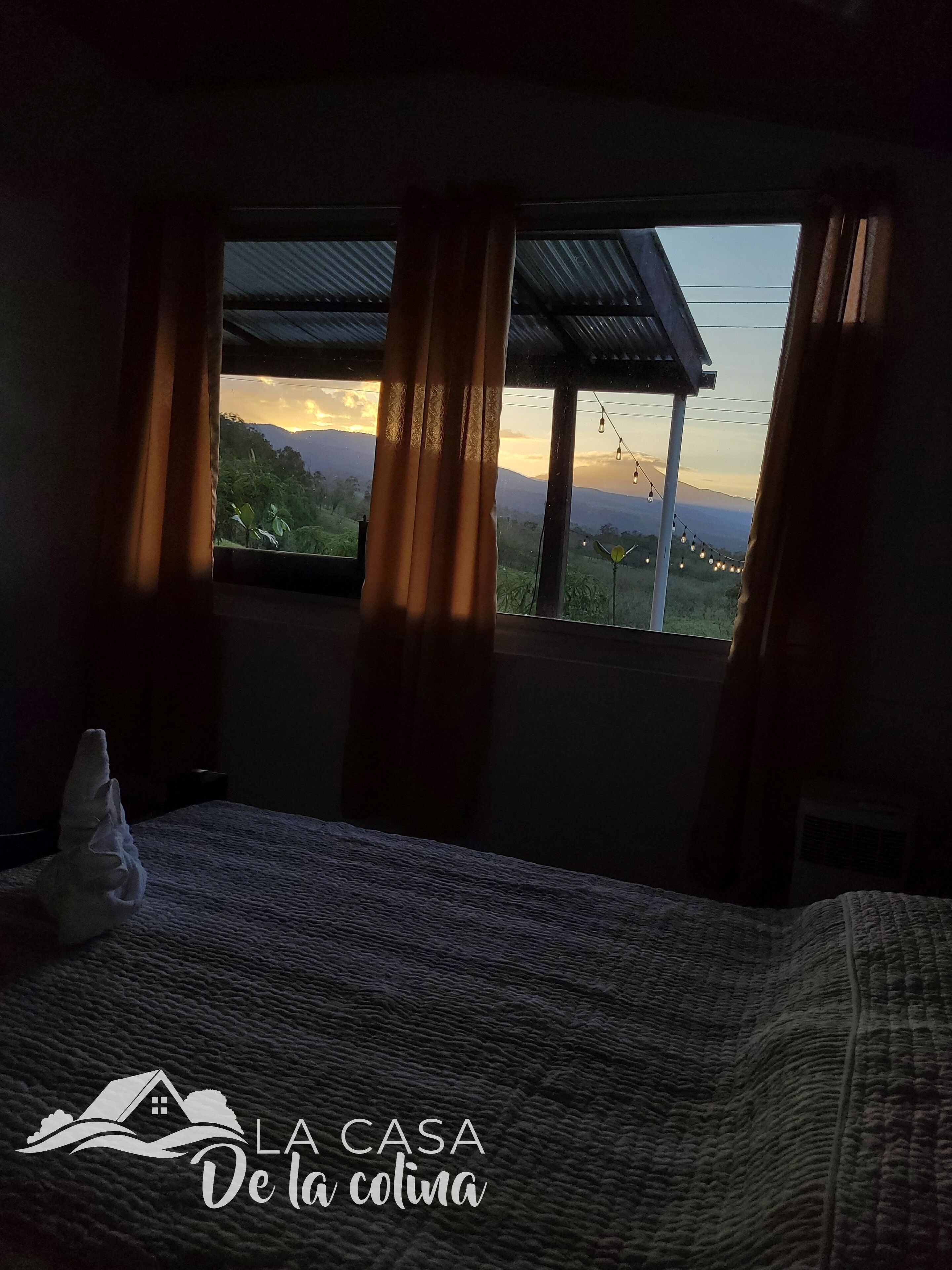 A bedroom with a bed, curtains, and a view of a sunset over mountains.