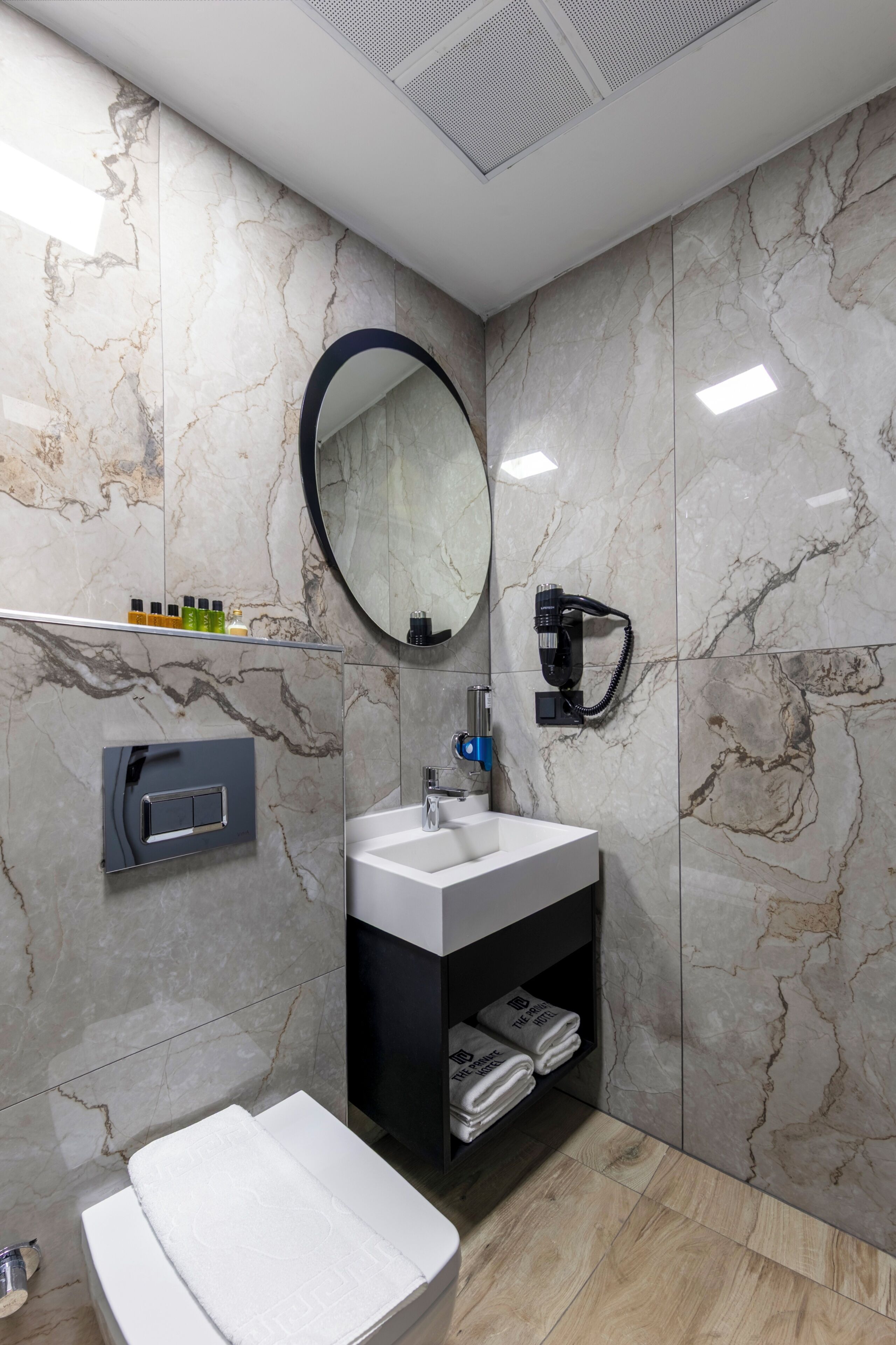 A modern bathroom with a marble wall, a round mirror, a sink, a towel rack, and a hairdryer.