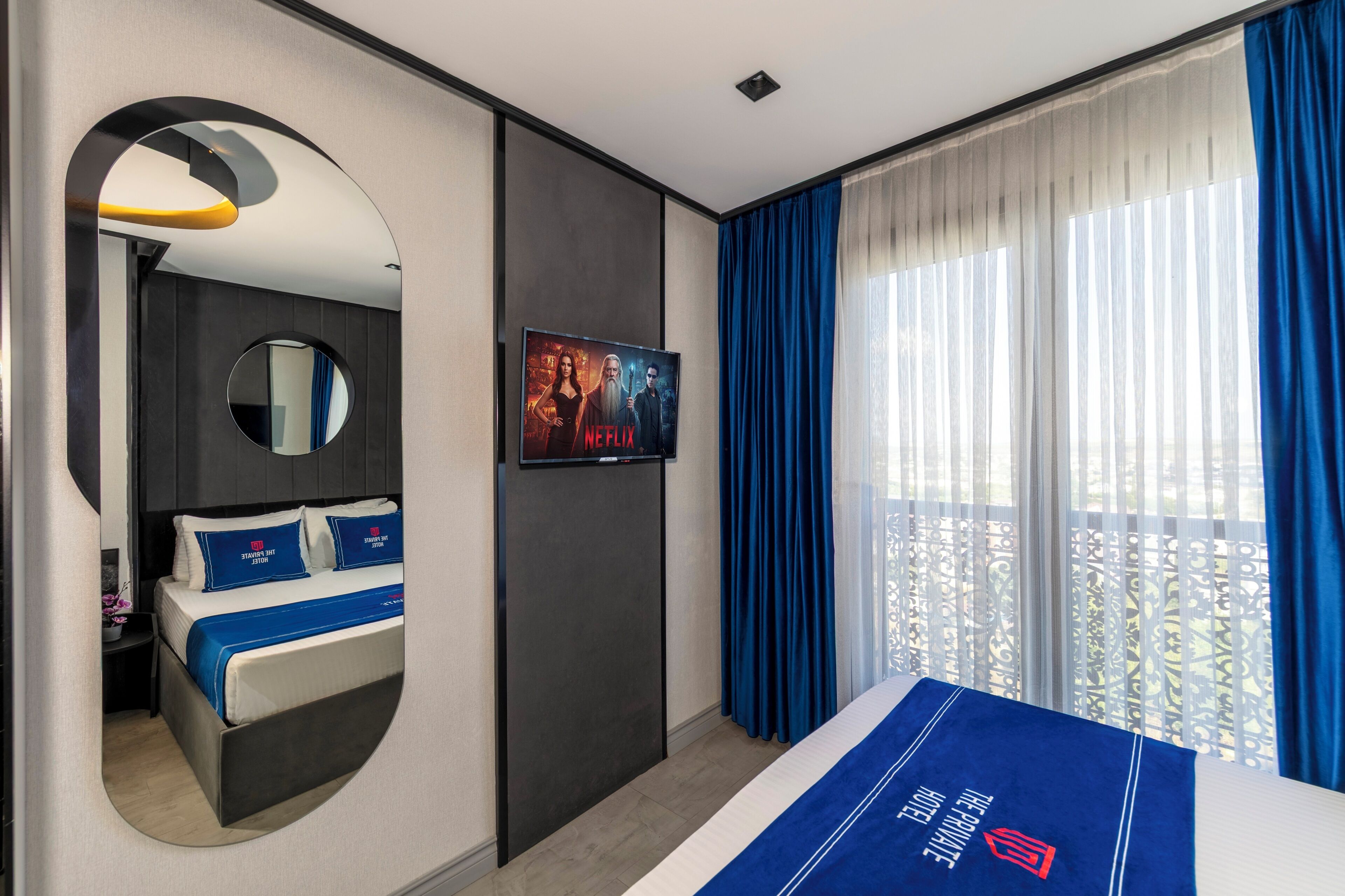 Una habitación de hotel moderna con cama, televisor, espejo y un amplio ventanal con cortinas.
