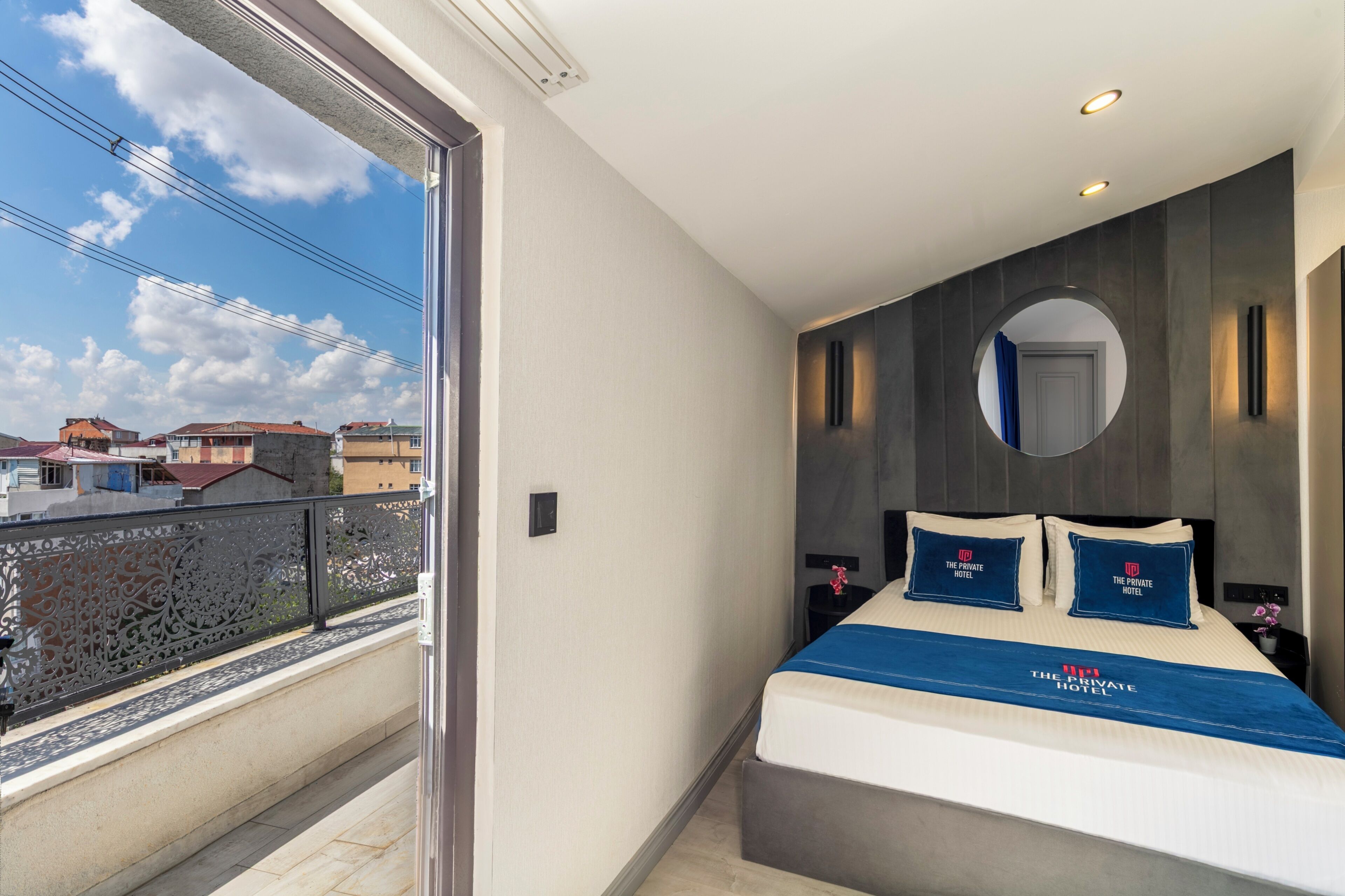 Een hotelkamer met balkon, een bed met beddeken van het merk, een ronde spiegel en een nachtkastje met lamp.