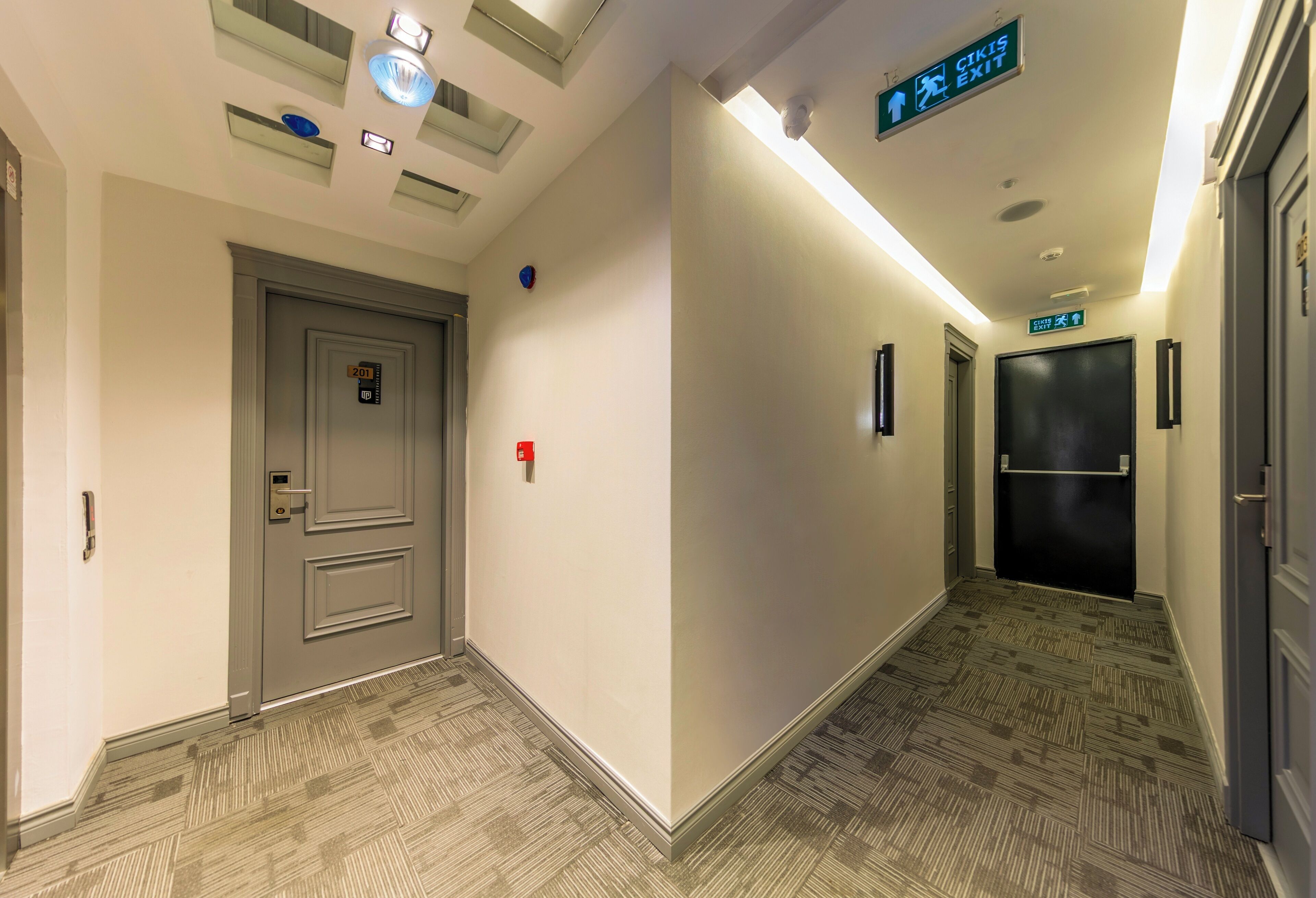 A hotel corridor with a grey door, a sign indicating the exit, and a patterned carpet.