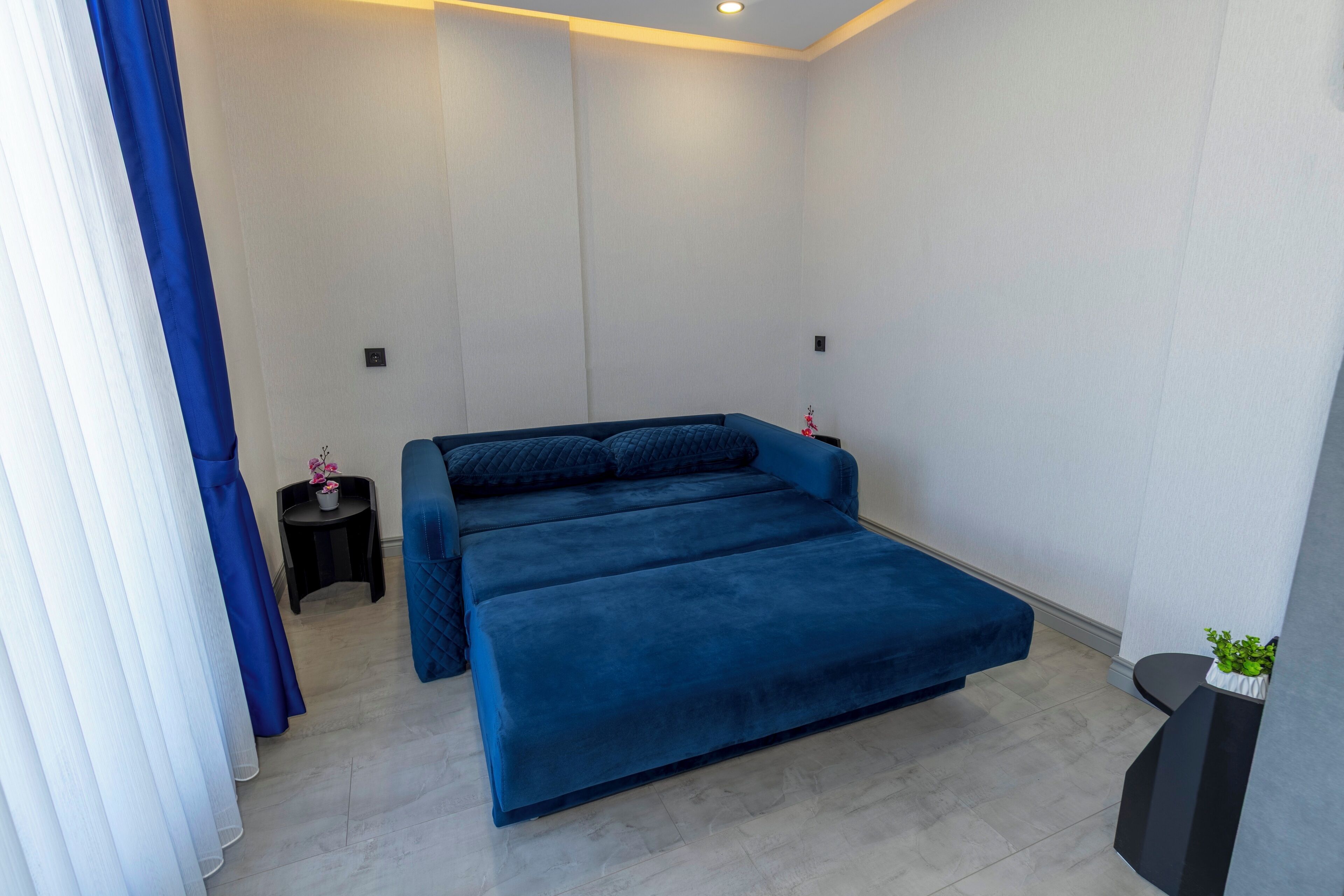 A modern bedroom with a large bed, a small bedside table with a plant, and a window with blue curtains.