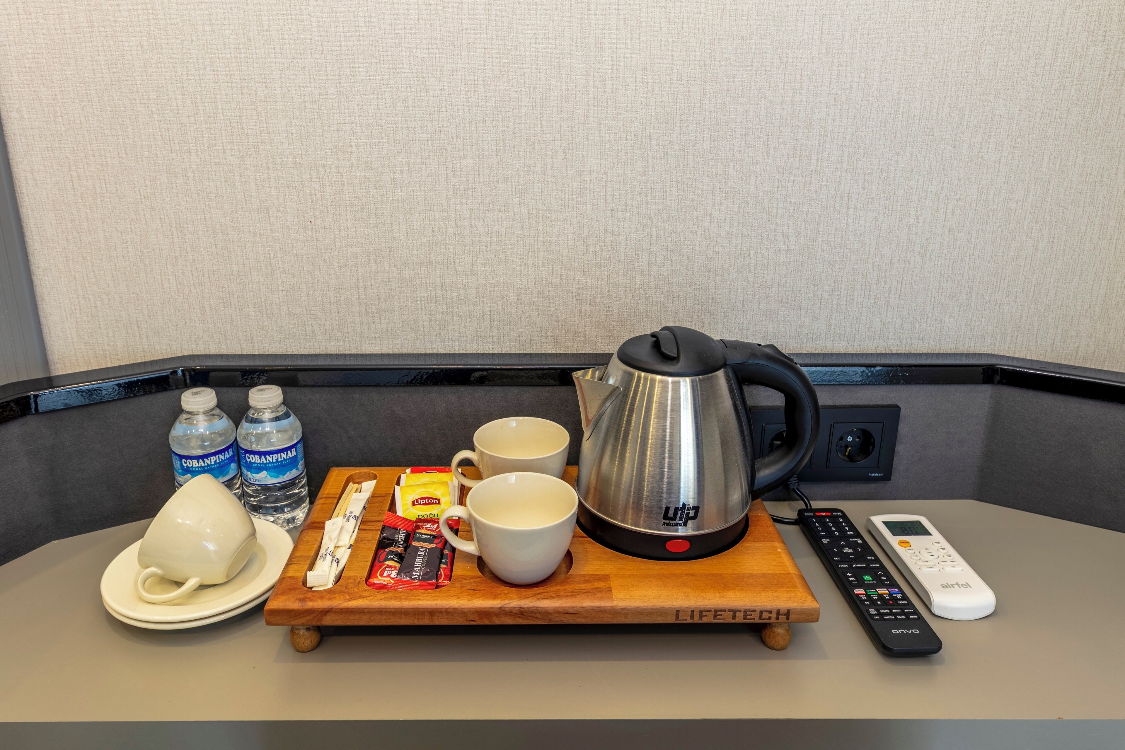 A breakfast setup with a kettle, two cups, a plate, a tray, two bottles of water, a remote control, and a mobile phone.