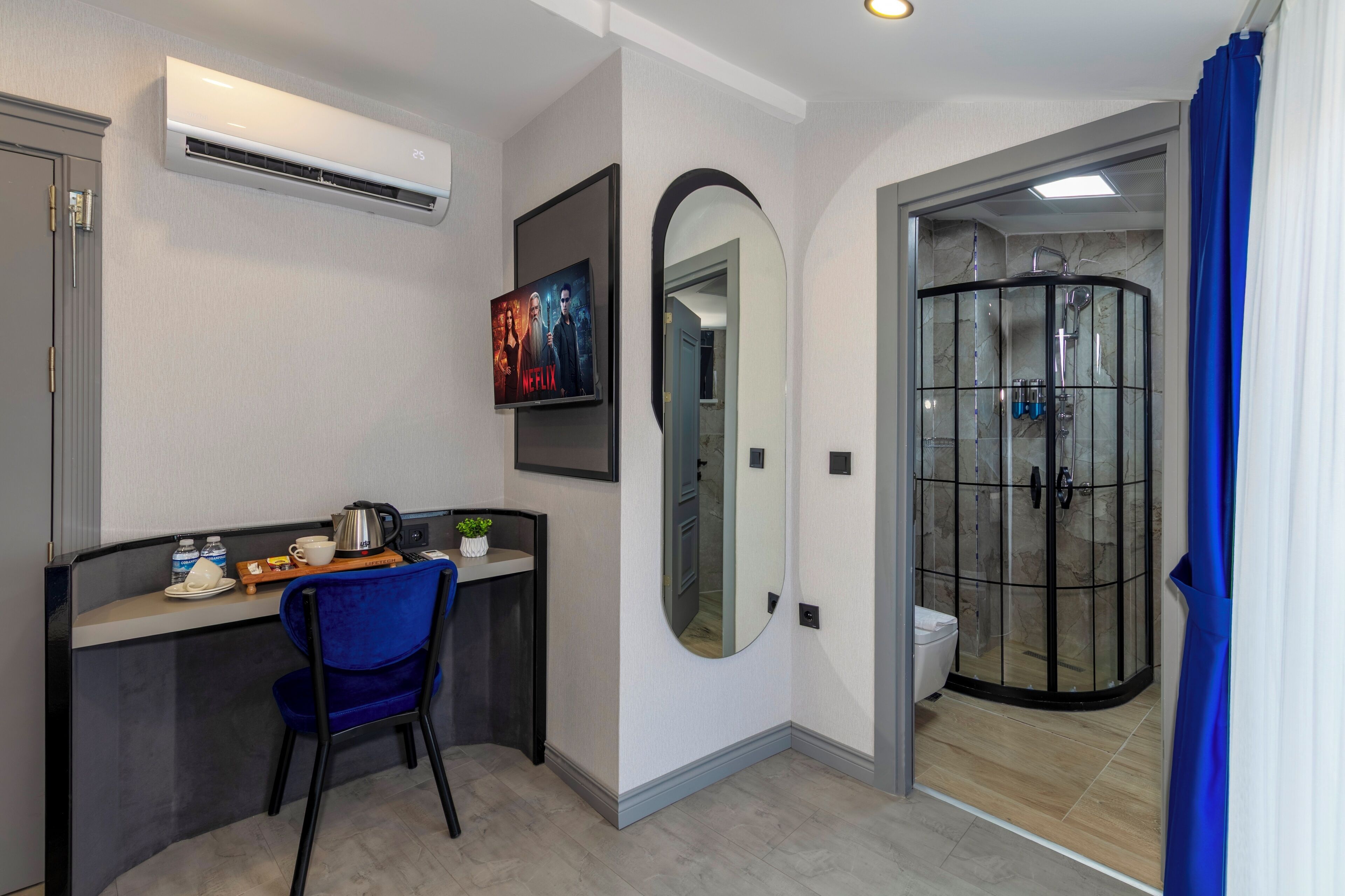 A hotel room with a desk, chair, air conditioning unit, TV, mirror, and a glass-enclosed shower area.