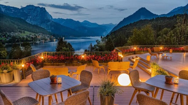 An outdoor seating area with tables and chairs, surrounded by potted plants and flowers, overlooking a lake and mountains. - Appartment mit Balkon und Seeblick (Grundlsee)