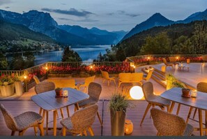An outdoor seating area with tables and chairs, surrounded by potted plants and flowers, overlooking a lake and mountains.