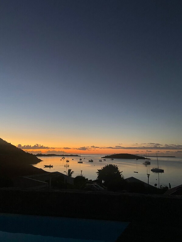 Un paysage côtier avec des bateaux au coucher du soleil, une eau calme et la silhouette d’un paysage vallonné.