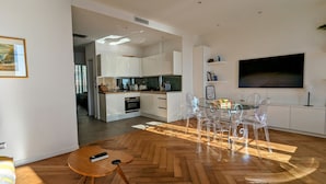 A modern kitchen with white cabinets, a dining area with clear chairs, and a large TV on the wall. - Happyfew Palazzo del sol (Nice)