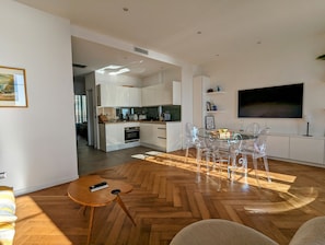 A modern kitchen with white cabinets, a dining area with clear chairs, and a large TV on the wall.