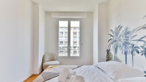 A hotel room with a bed, a chair, a window with a view of a building, and a wall with a palm tree mural. - Happyfew Palazzo del sol (Nice)