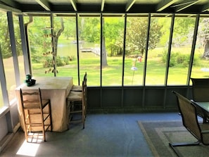 Une véranda avec une table et des chaises, donnant sur un étang et la verdure environnante.