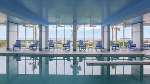 Un espace piscine avec des chaises et des tables bleues, de grandes fenêtres et une vue sur l’océan.