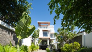 Une maison moderne à deux étages, dotée d’une entrée centrale, entourée d’une végétation tropicale luxuriante et surplombée d’un ciel bleu azur.