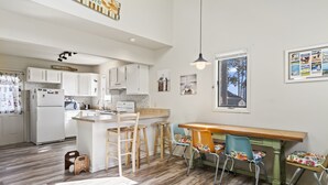 A kitchen with white cabinets, a central island with stools, a wooden dining table with chairs, and a hanging light fixture. - Channel View Charmer by Seaside Vacations & Sales (Chincoteague Island)