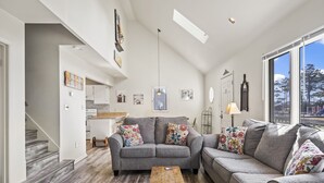 A modern living room with a grey sofa set, a wooden coffee table, and a view of a park through large windows. - Channel View Charmer by Seaside Vacations & Sales (Chincoteague Island)