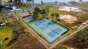 An aerial view of a tennis court complex with multiple courts, a parking area, and a body of water in the background. - Channel View Charmer by Seaside Vacations & Sales (Chincoteague Island)