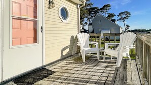 A wooden deck with two white Adirondack chairs, a door with a pink panel, and a house in the background. - Channel View Charmer by Seaside Vacations & Sales (Chincoteague Island)