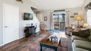 Un salon spacieux doté d’un plancher en bois, d’un canapé, d’une table basse et d’une cheminée. Une porte donne accès à un balcon d’où l’on peut admirer la vue sur les arbres.